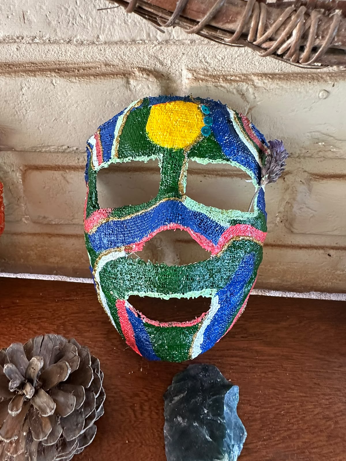 Máscara de gesso pintada em azul, verde e dourado, pendurada na parede